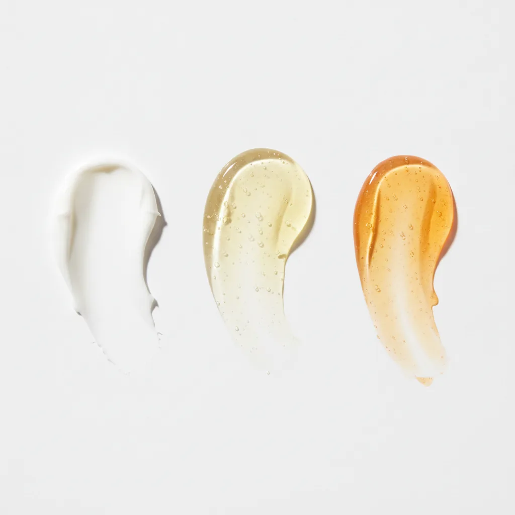 Een close-up van drie verschillende cosmetische texturen naast elkaar, van een witte crème tot een gele gel en een oranje serum, die de verschillende soorten retinoïden symboliseren.